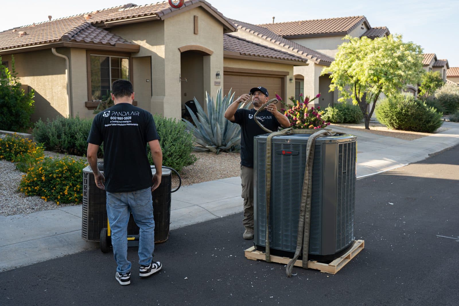 A new condensing unit strapped on a pallet and the old unit on a dolly in front of a house, with two technicians